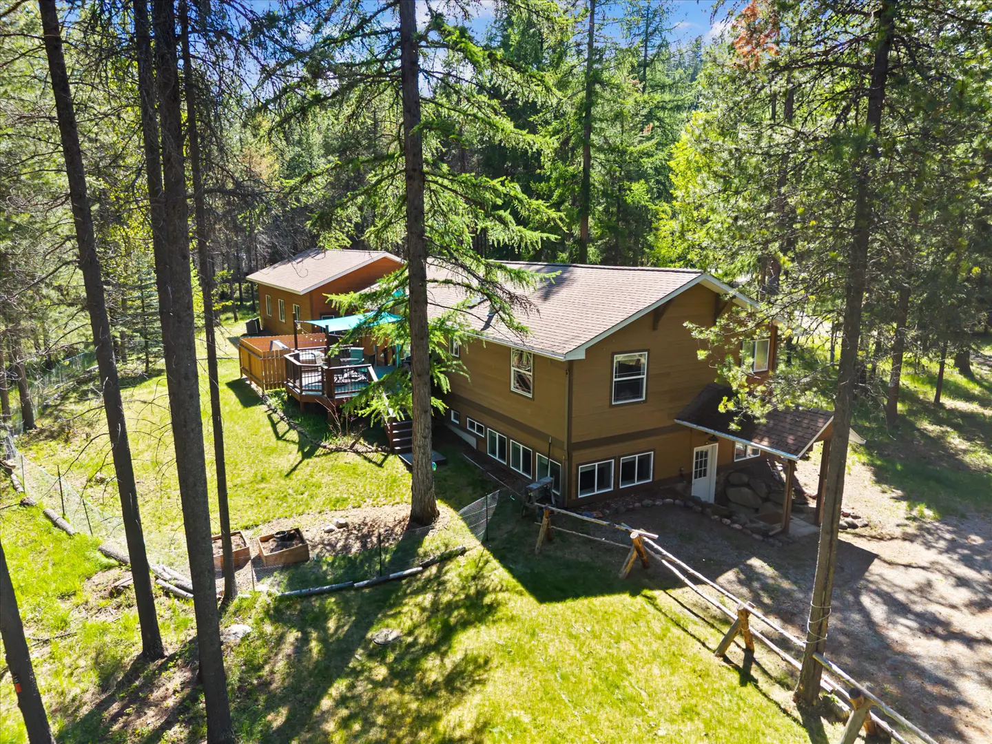 Aerial view of a brown two-story house with a deck and a blue awning, surrounded by tall trees and green grass.