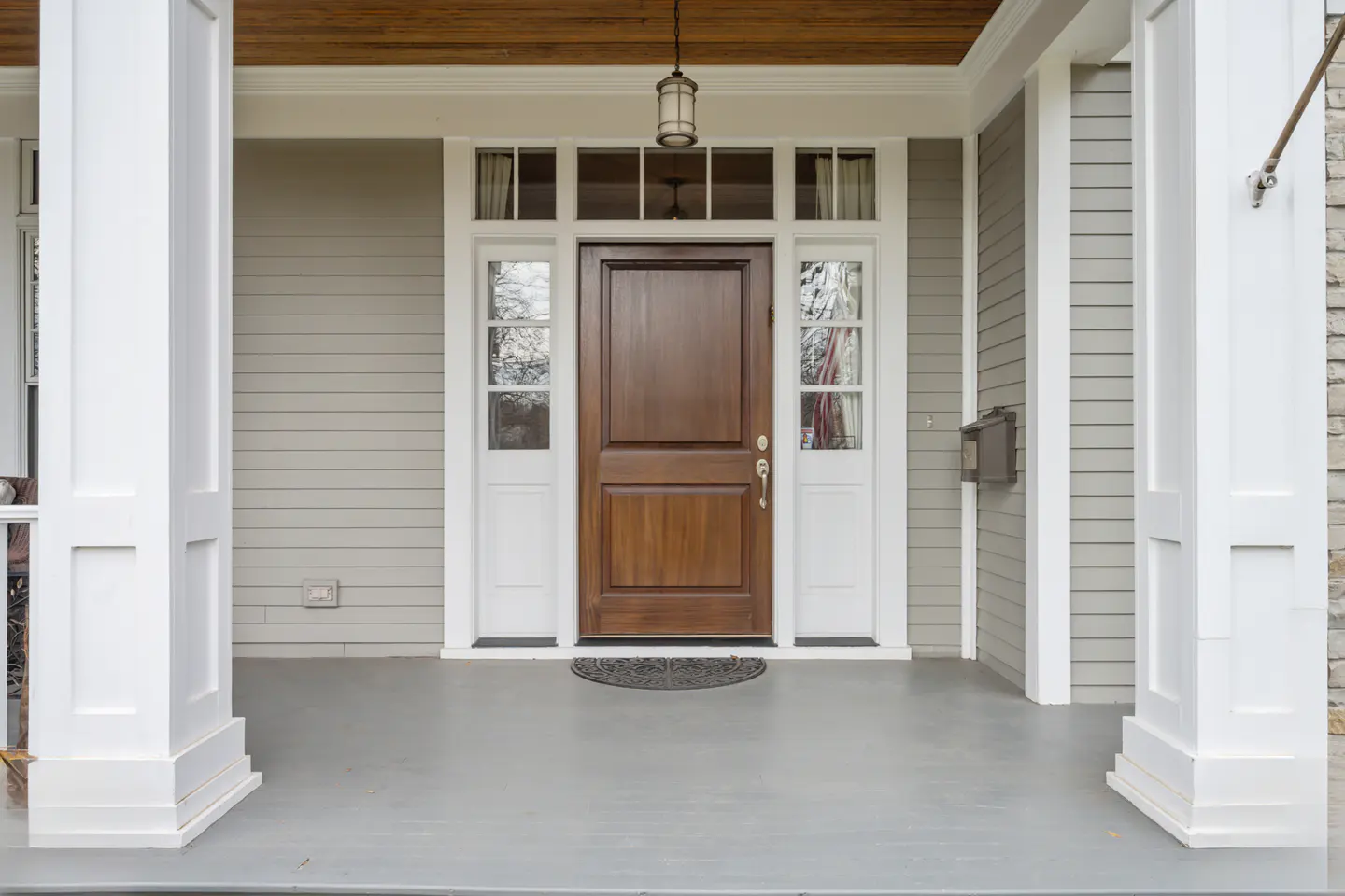 Front porch of a house with a dark wood door, white trim, and gray siding. White columns frame the entrance. A light fixture hangs above the door.