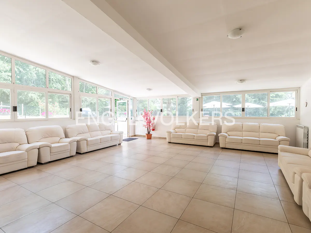 Spacious, sunlit lounge with white leather sofas, tiled floors, and large windows overlooking a green garden.