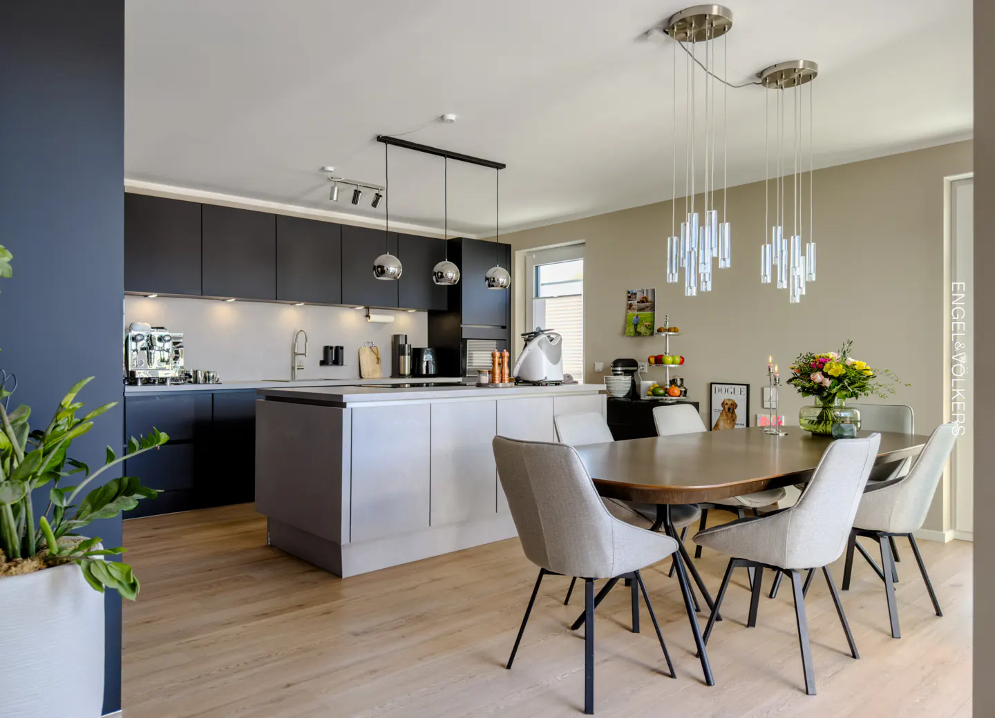 A modern kitchen with black cabinets, a gray island, and a round wooden dining table with six gray chairs. Cylindrical pendant lights hang above the table.