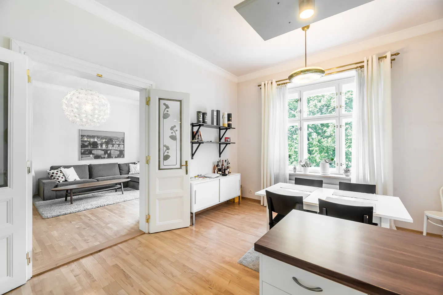 Bright, airy room with hardwood floors, white walls, and open doorway to a living room with a gray sofa. Dining table near a window.