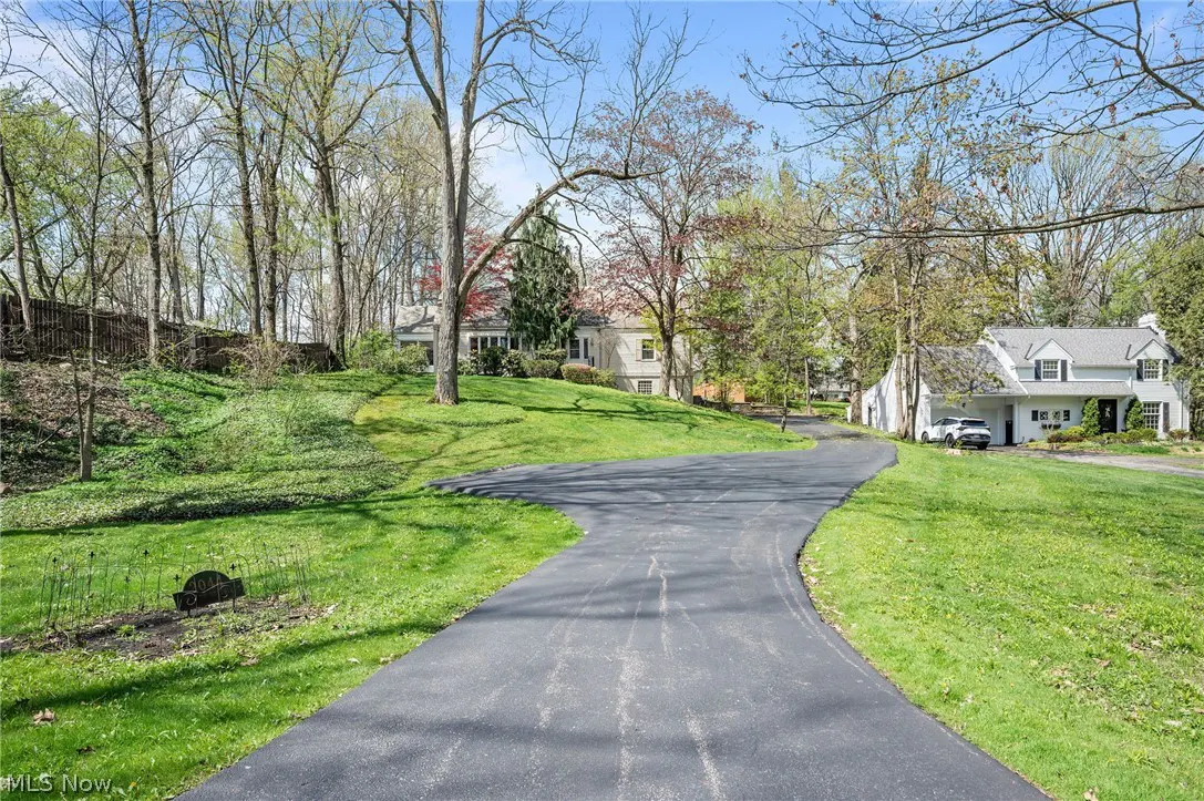 A long, black driveway leads to a white house with a black roof, surrounded by green grass and trees.