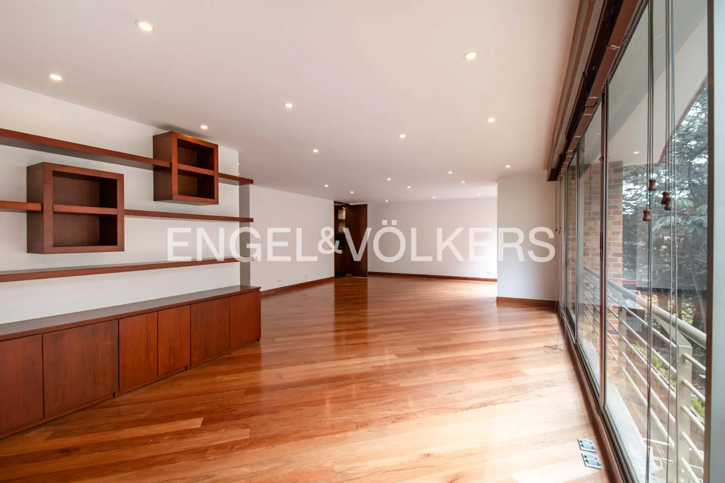 Bright, empty living room with hardwood floors, white walls, and floor-to-ceiling windows. Brown shelving and cabinets line the left wall.