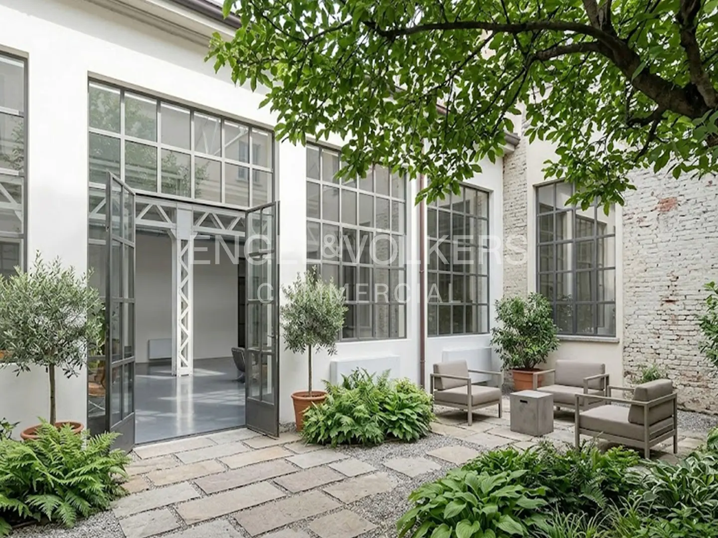 Outdoor patio with stone pavers, gravel, and greenery. Gray chairs and a table sit near a white building with large windows and open doors.