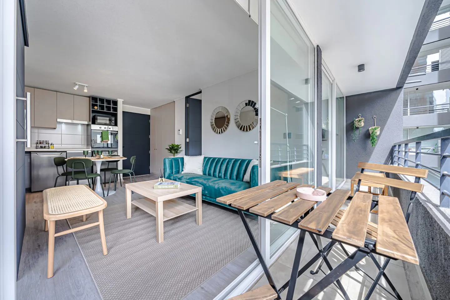 Modern apartment interior with a teal velvet sofa, light wood coffee table, dining area, and a balcony with wooden furniture.