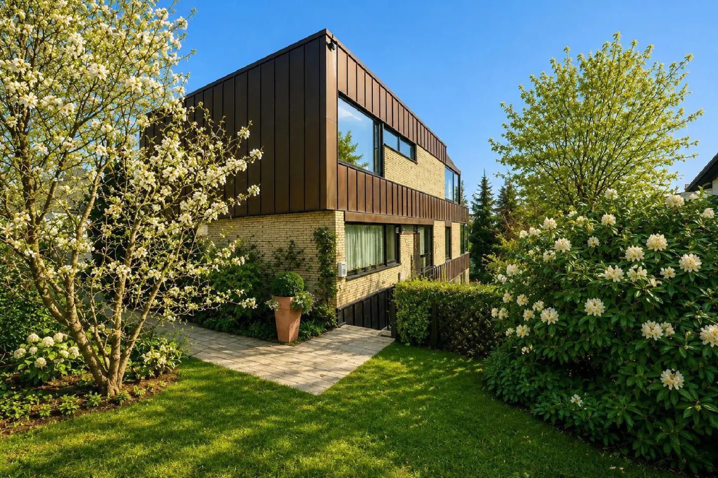 A modern two-story house with a brown metal roof and light brick exterior, surrounded by lush green landscaping and flowering trees.