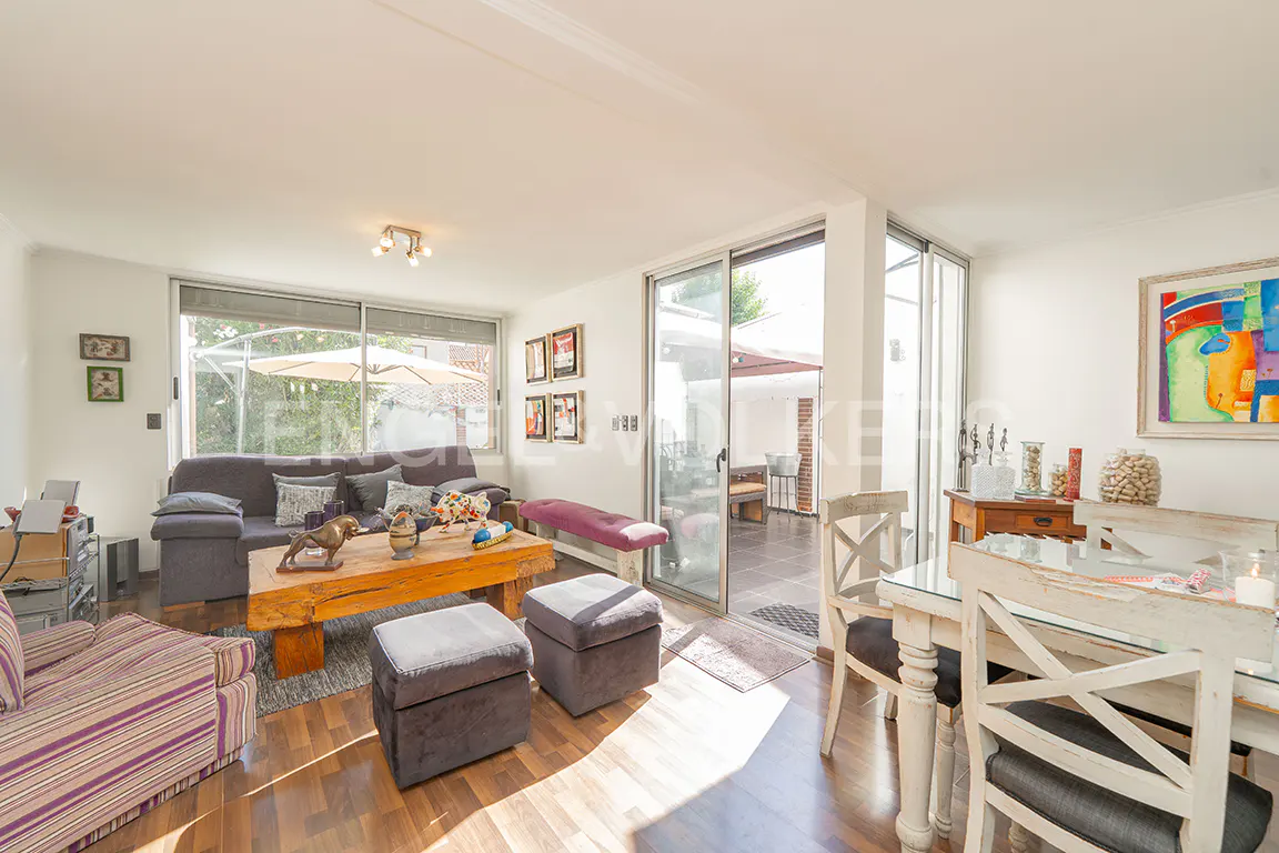 Bright living room with a grey sofa, rustic wooden coffee table, two grey ottomans, and a dining area with white chairs.