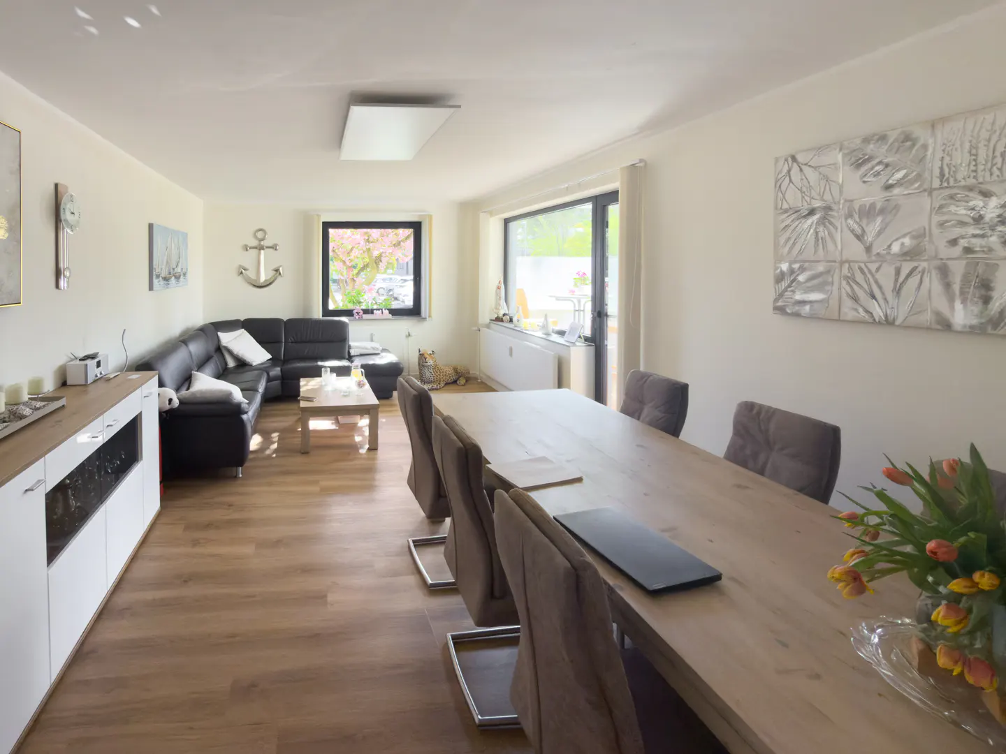 A bright, open-concept living and dining area with a black leather sofa, wood floors, and a long dining table with gray chairs. Tulips in a vase sit on the table.