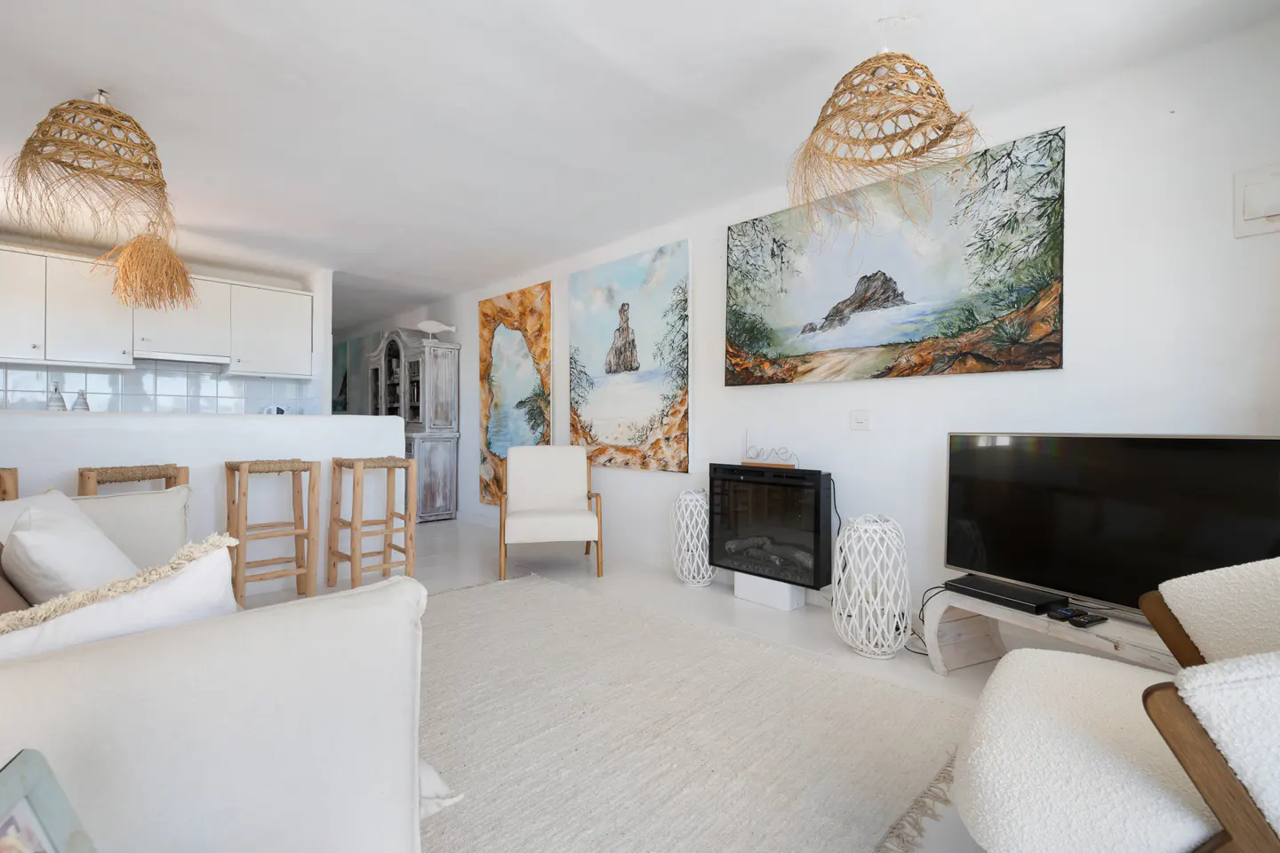 Bright, all-white living room with straw pendant lights, beach-themed art, and a modern fireplace. Kitchen area visible in the background.