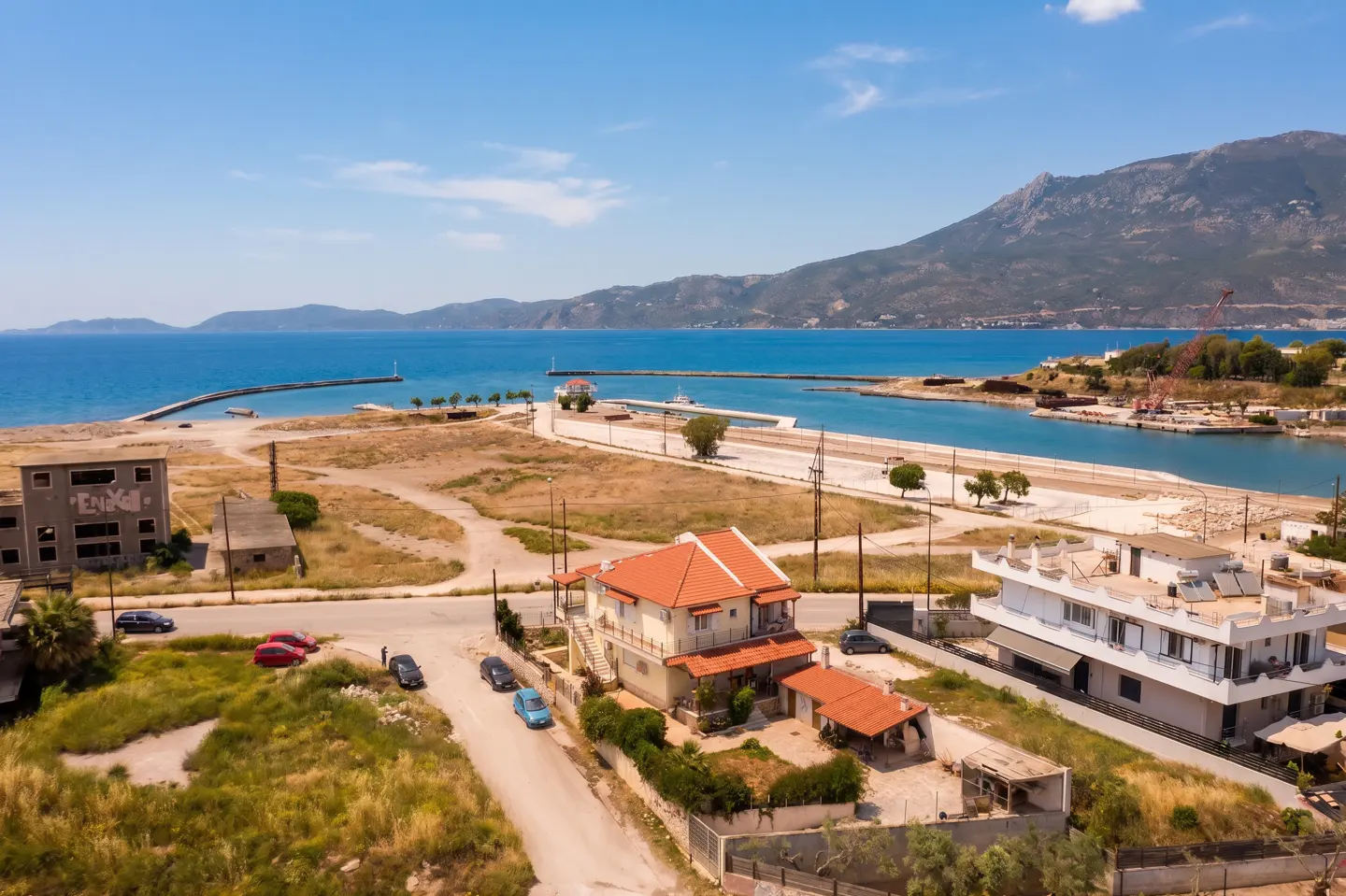 Aerial view of coastal real estate. Houses with red tile roofs near a harbor with blue water and mountains in the background.