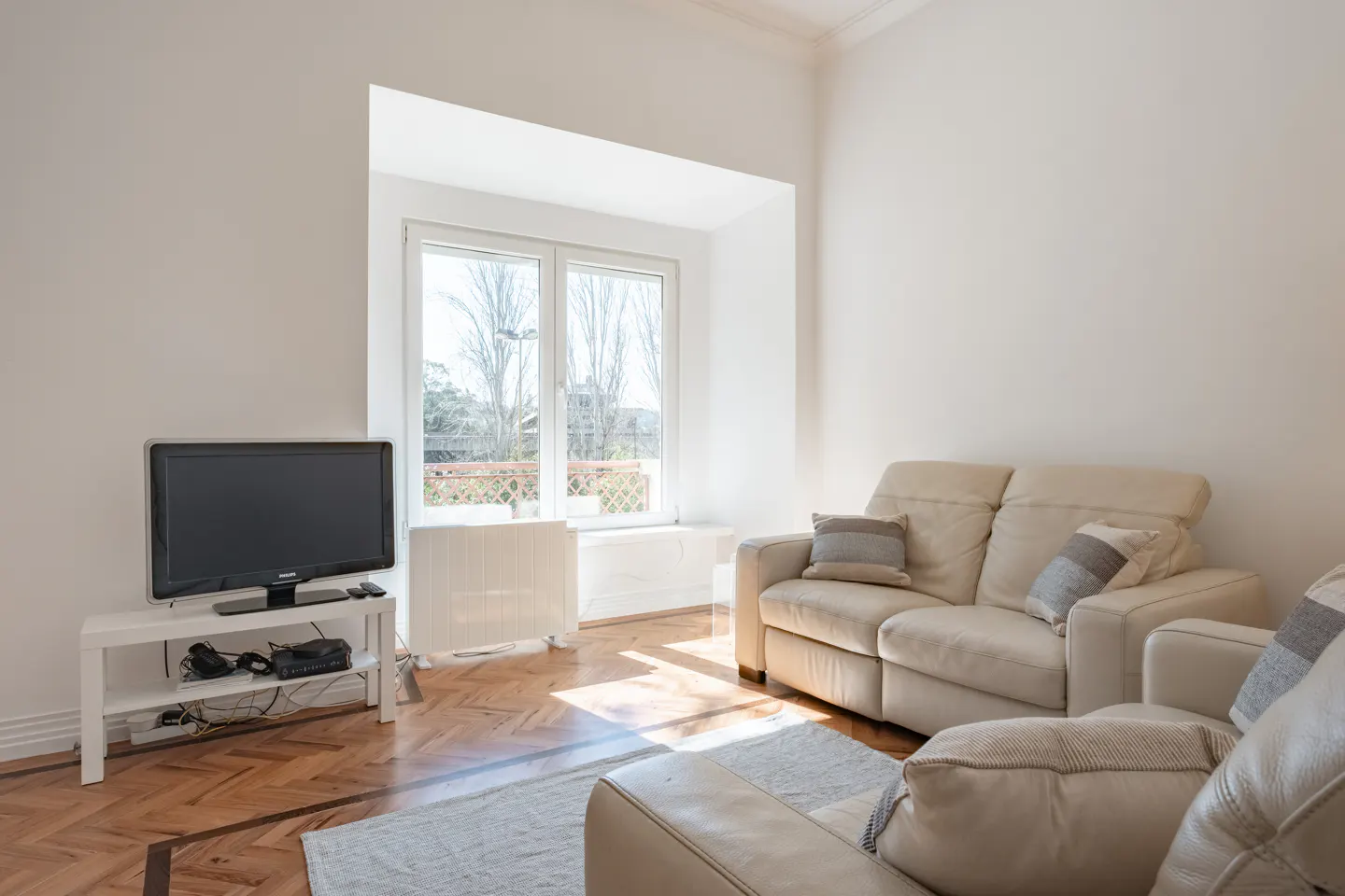 Bright living room with white walls, parquet floor, and beige leather sofas. A TV sits on a white stand near a window with a view.