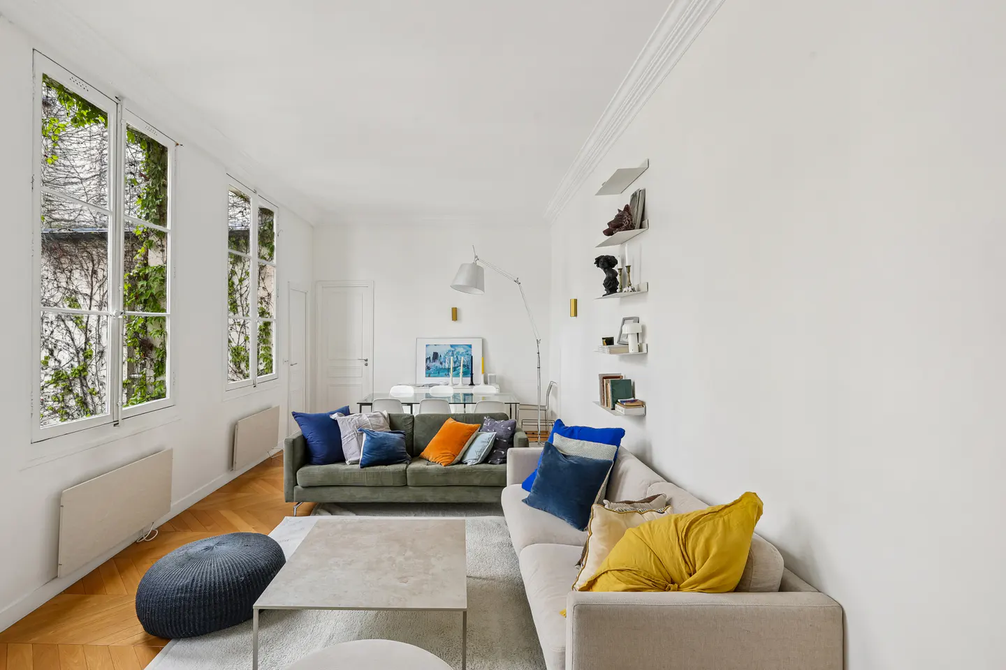 Bright living room with hardwood floors, white walls, and large windows with green vines. Two sofas with colorful pillows face a coffee table. Shelves adorn the wall.
