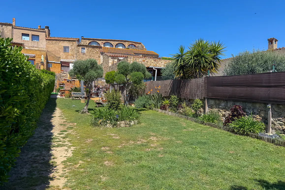 A sunny backyard with green grass, trees, and a stone wall with a brown fence. Old stone buildings are in the background.