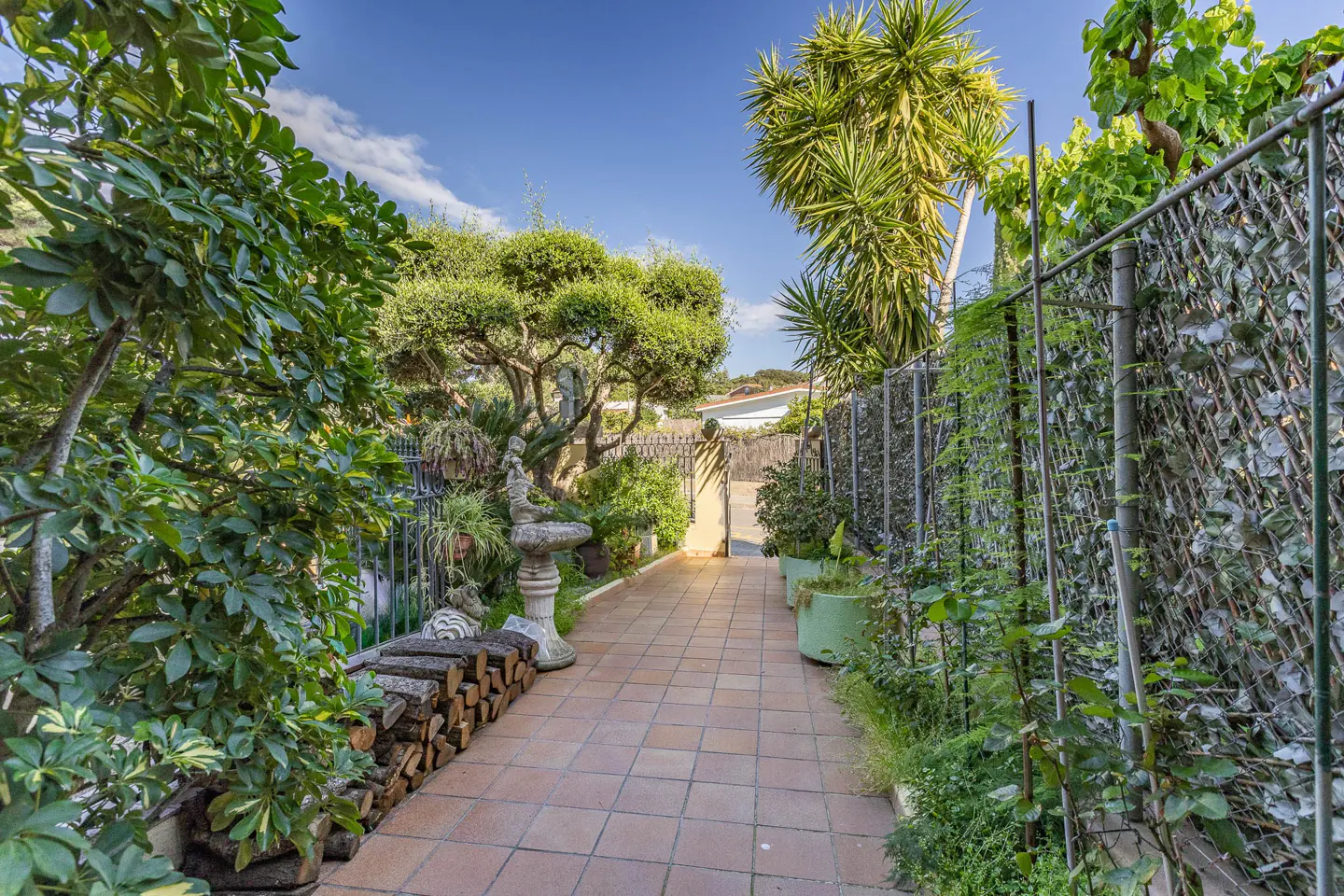 A garden path with terracotta tiles leads to a gate, lined with lush greenery, a stone fountain, and stacked firewood.
