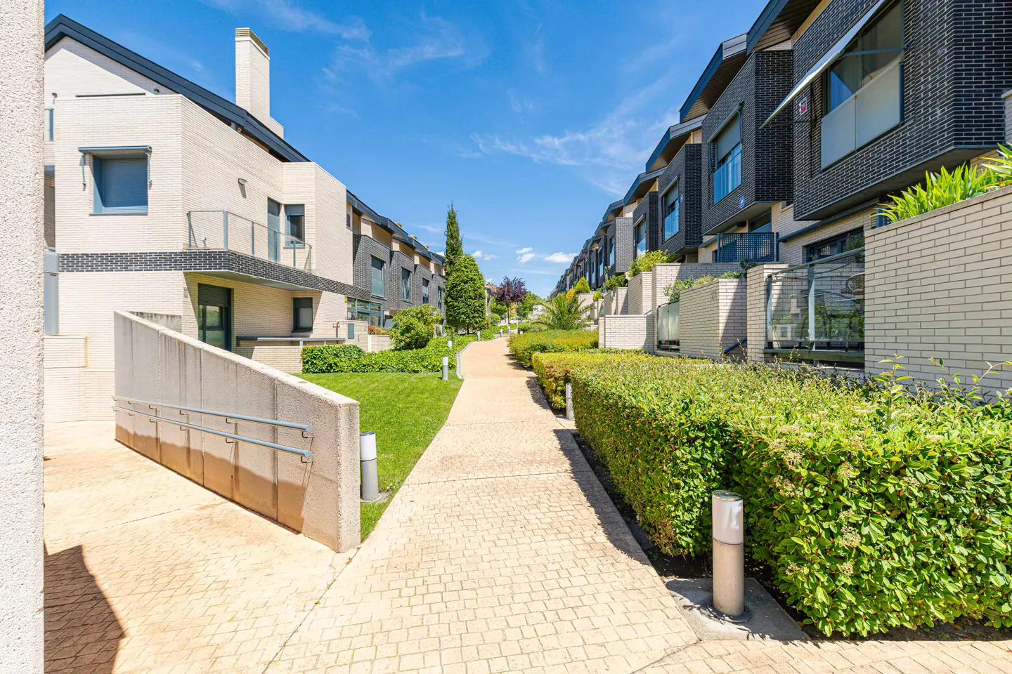 A row of modern townhouses with brick facades and manicured green hedges under a bright blue sky.
