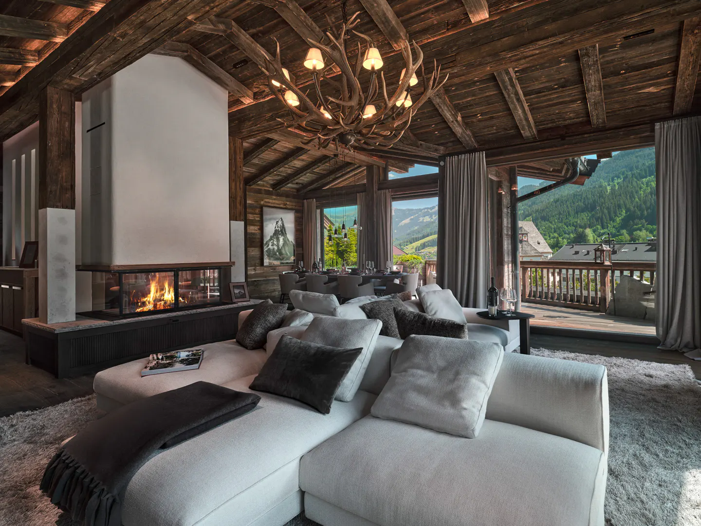 Luxury mountain chalet living room with a white sectional sofa, antler chandelier, and fireplace overlooking green hills.