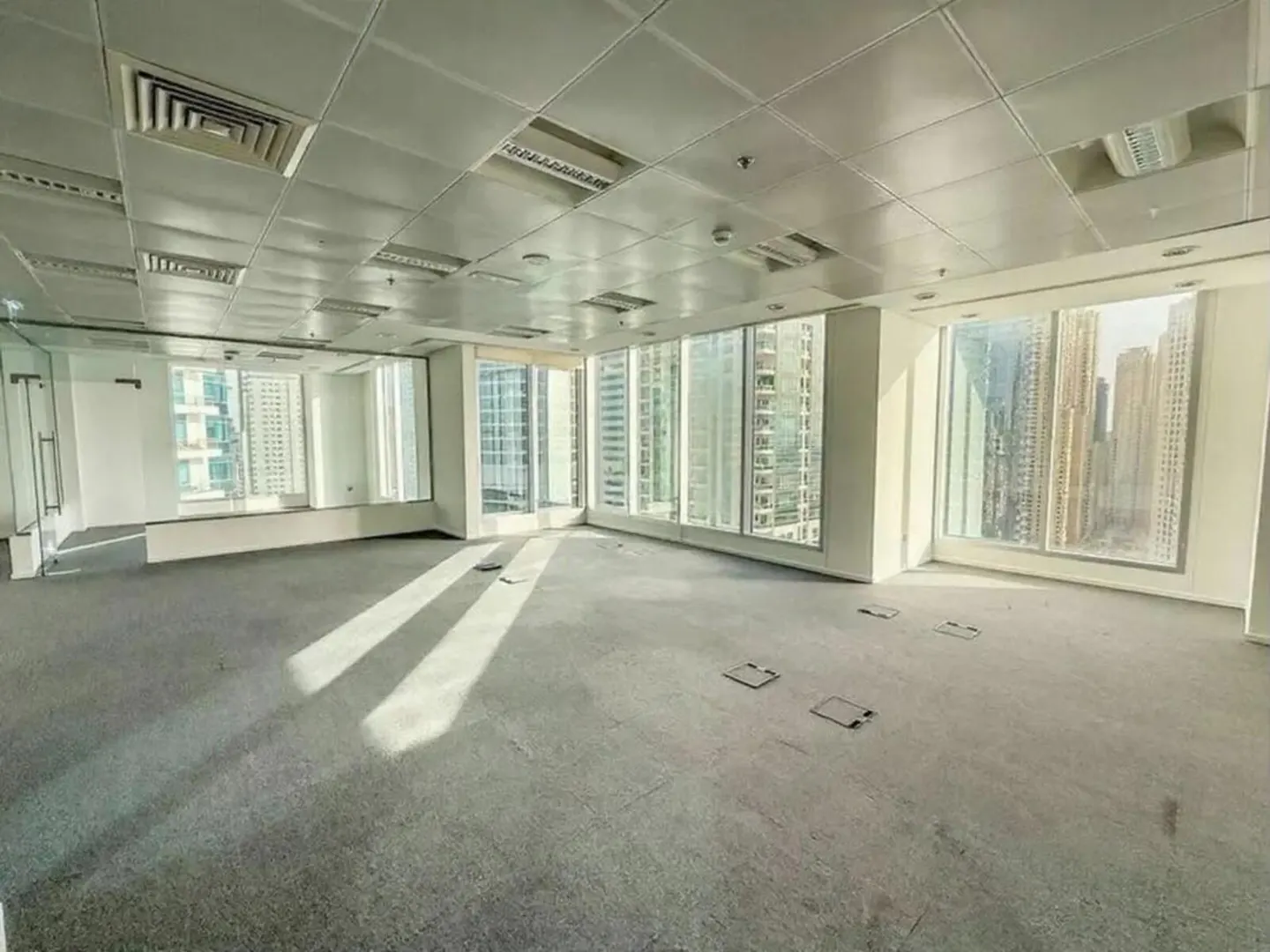 Empty office space with gray carpet, white walls, and large windows showing a city skyline. The ceiling has recessed lighting and vents.