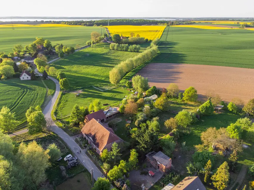 Aerial view of a rural landscape with fields of yellow flowers, green crops, and a farmhouse with a red tile roof.