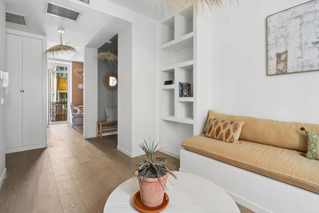 Bright apartment interior with wood floors, white walls, and built-in shelves. A tan sofa with patterned pillows sits near a round white table with a potted plant. A balcony is visible through an open doorway.