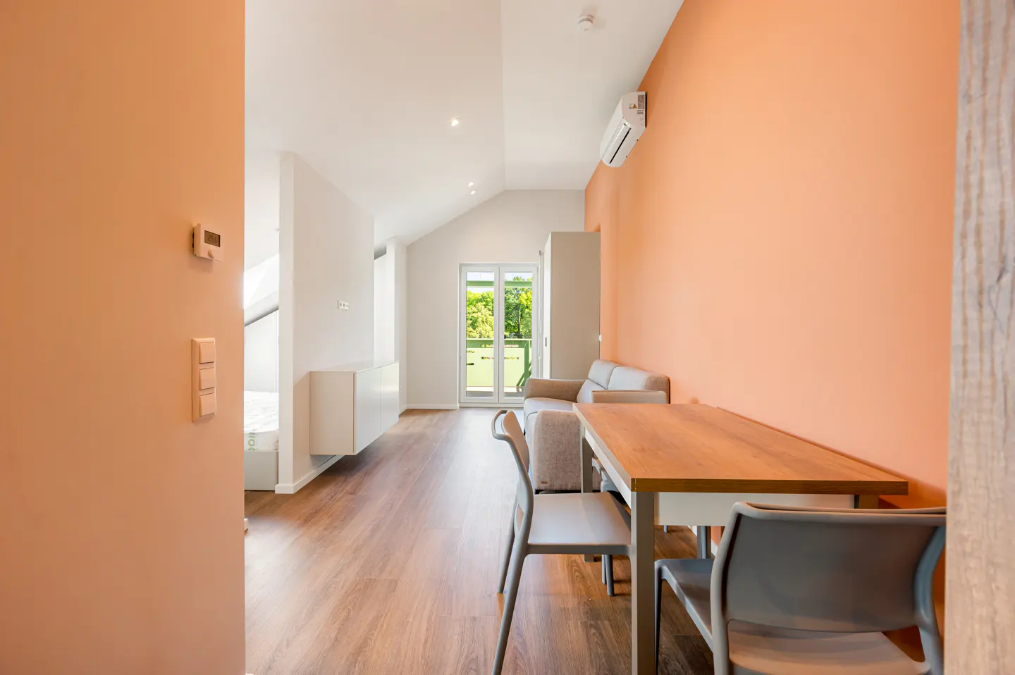 A bright apartment interior with peach walls, wood floors, and modern furniture. A dining table with chairs sits near a sofa, with a balcony visible in the background.