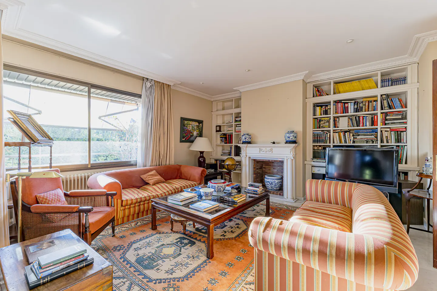 Bright living room with orange sofas, a fireplace, and a large bookshelf filled with books. A patterned rug covers the floor.