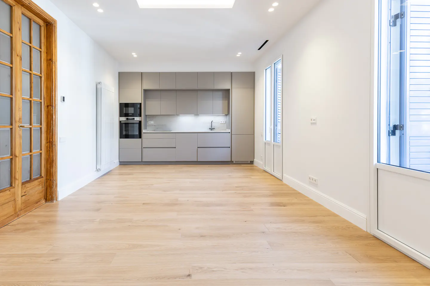Bright, empty room with light wood floors, white walls, and a gray kitchen. A wooden door is on the left and a window on the right.
