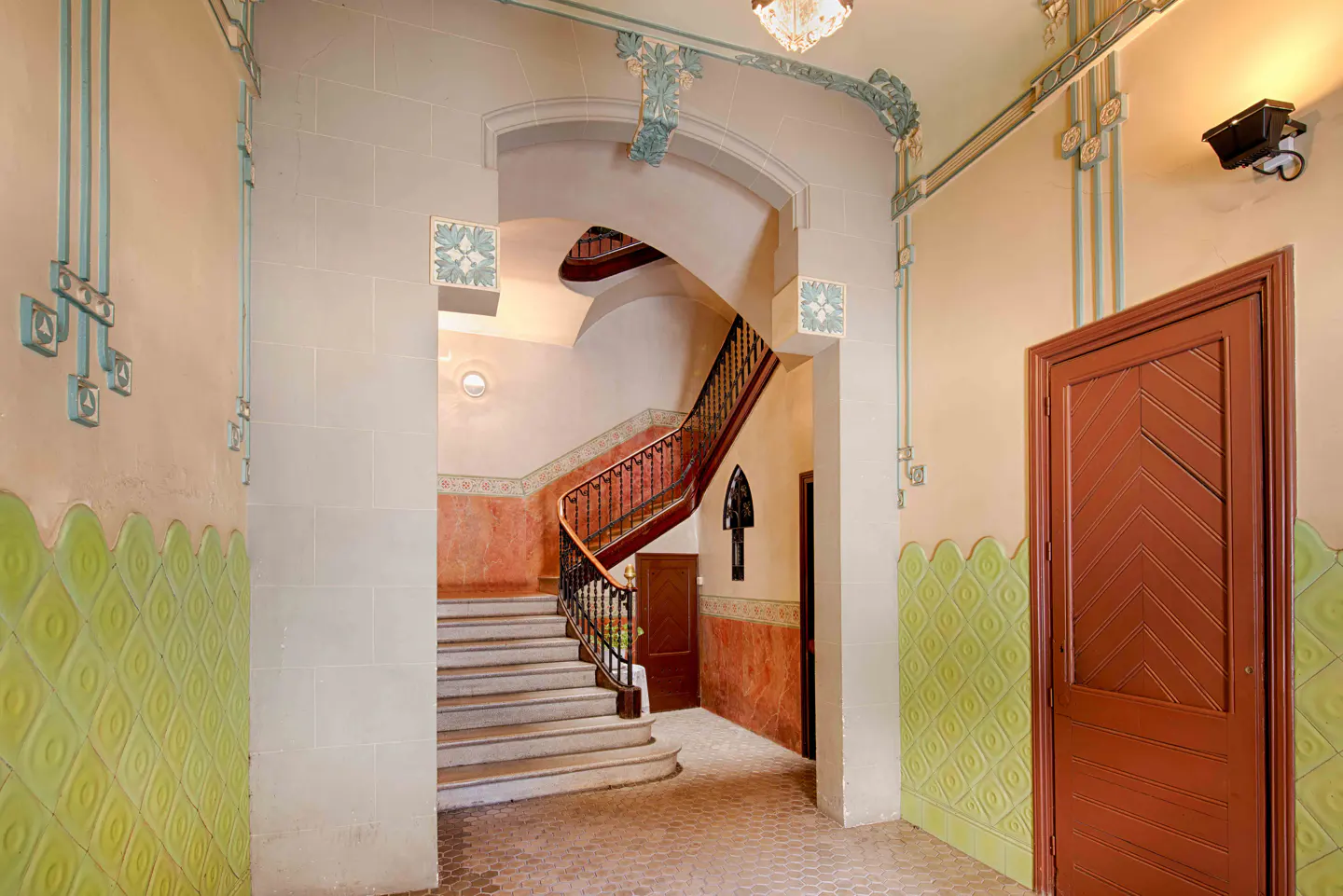 Interior view of a building's entrance hall with a staircase. Walls are decorated with green tiles and blue accents. A brown door is on the right.