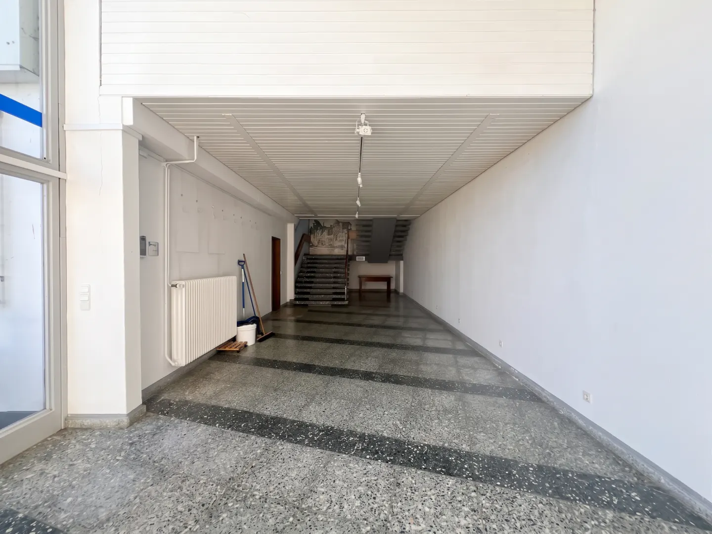 A long, empty hallway with white walls and a gray terrazzo floor. Stairs and a table are at the end of the hall.
