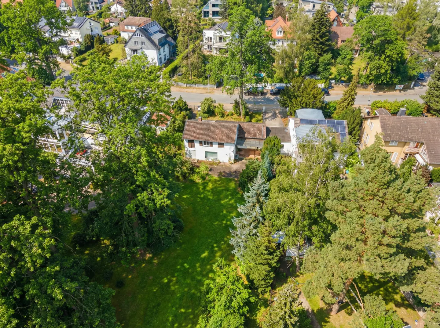 Aerial view of a white house with a brown roof, surrounded by green trees and a lawn. Other houses are visible in the background.