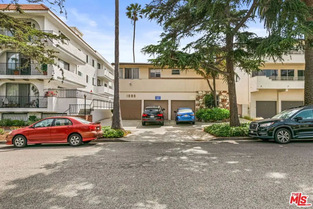 Street view of a two-story apartment building with three garage doors. Cars are parked in front and on the street. Palm trees and greenery surround the building.