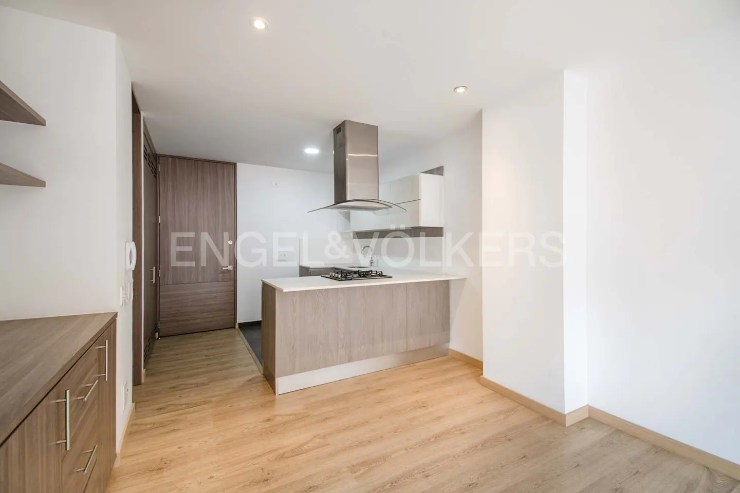 A modern kitchen with light wood floors, white walls, and a gray island with a white countertop. A stainless steel range hood hangs above the island.