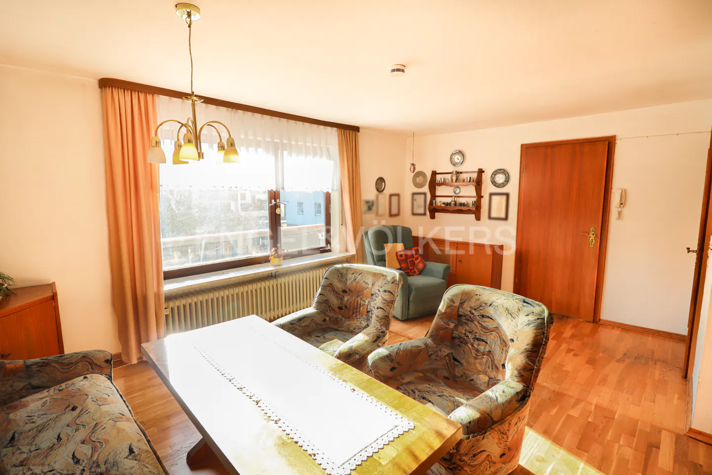 Living room with floral armchairs, a wooden table with a white runner, and a window with peach curtains.