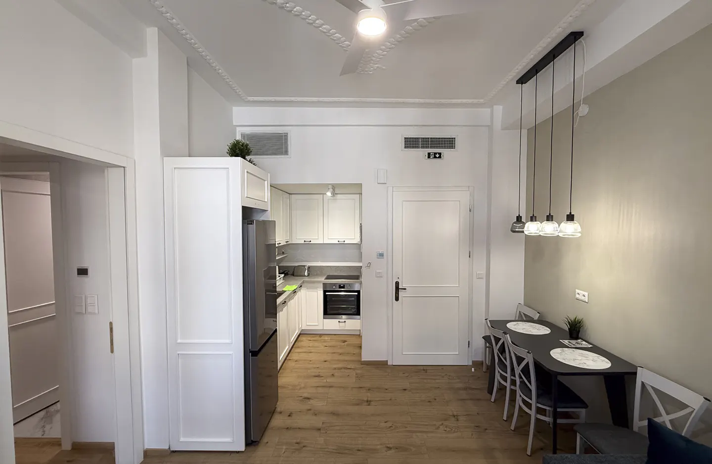 Bright, modern apartment interior with white walls, wood floors, and a dining table with four chairs. A white kitchen is visible in the background.