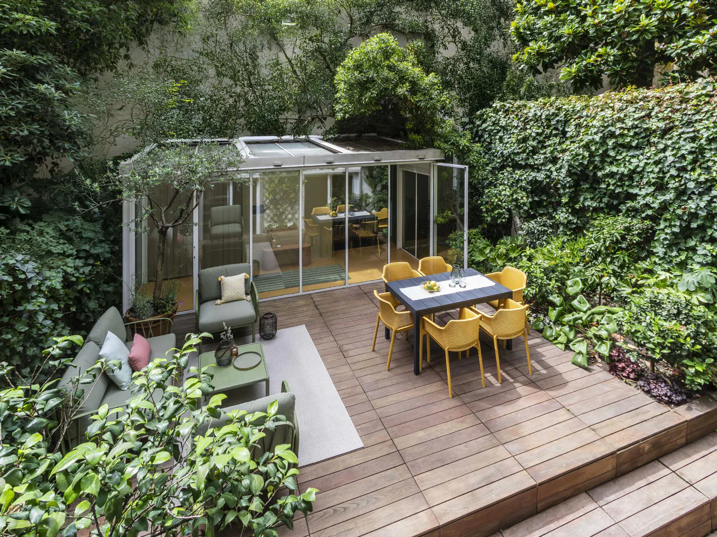 Outdoor patio with wood flooring, green sofa, yellow chairs around a black table, and a glass enclosed sunroom.