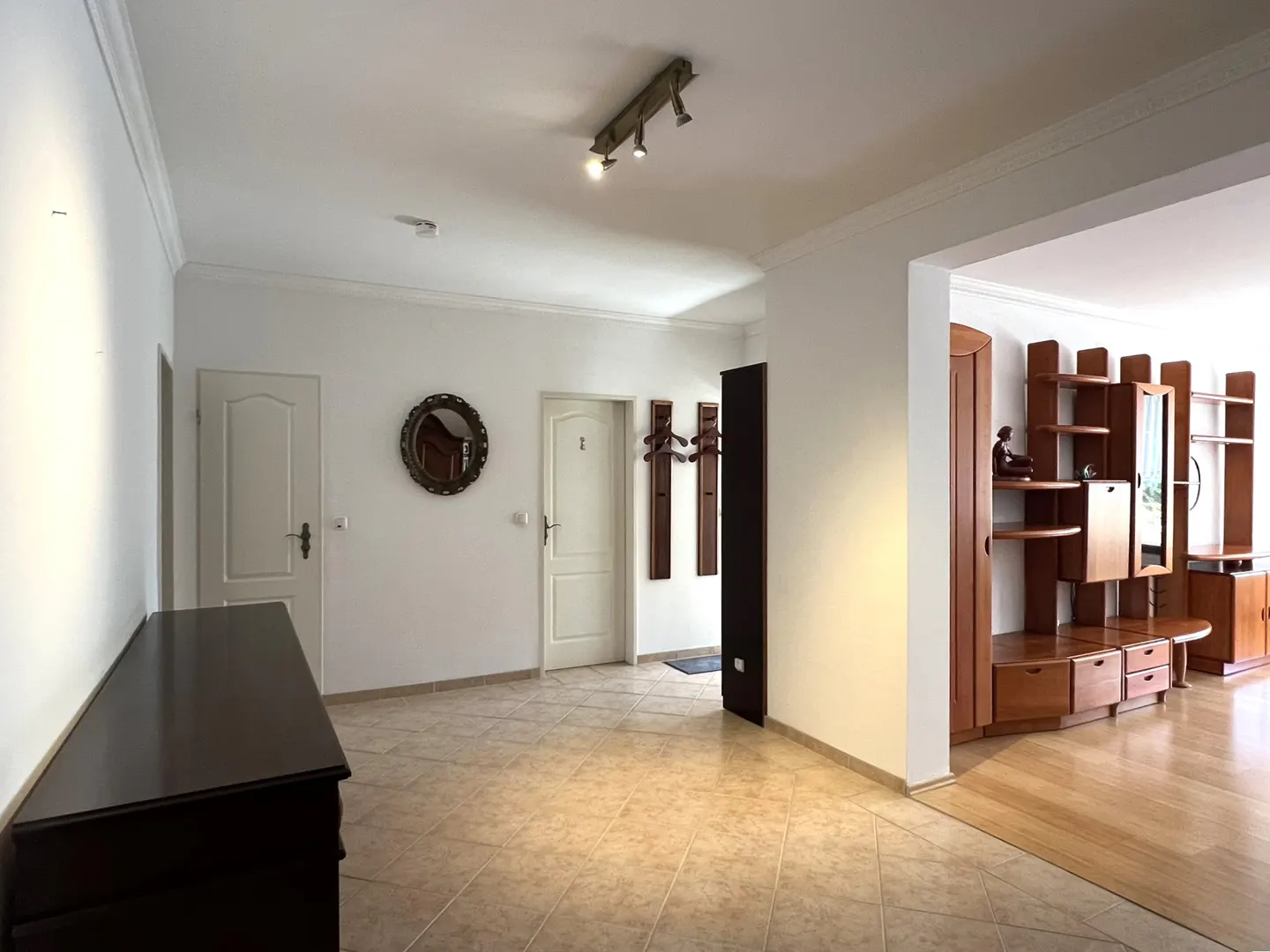A bright hallway with beige tile floors, white walls, and two white doors. A dark wood dresser sits to the left. A living room with wood furniture is visible.