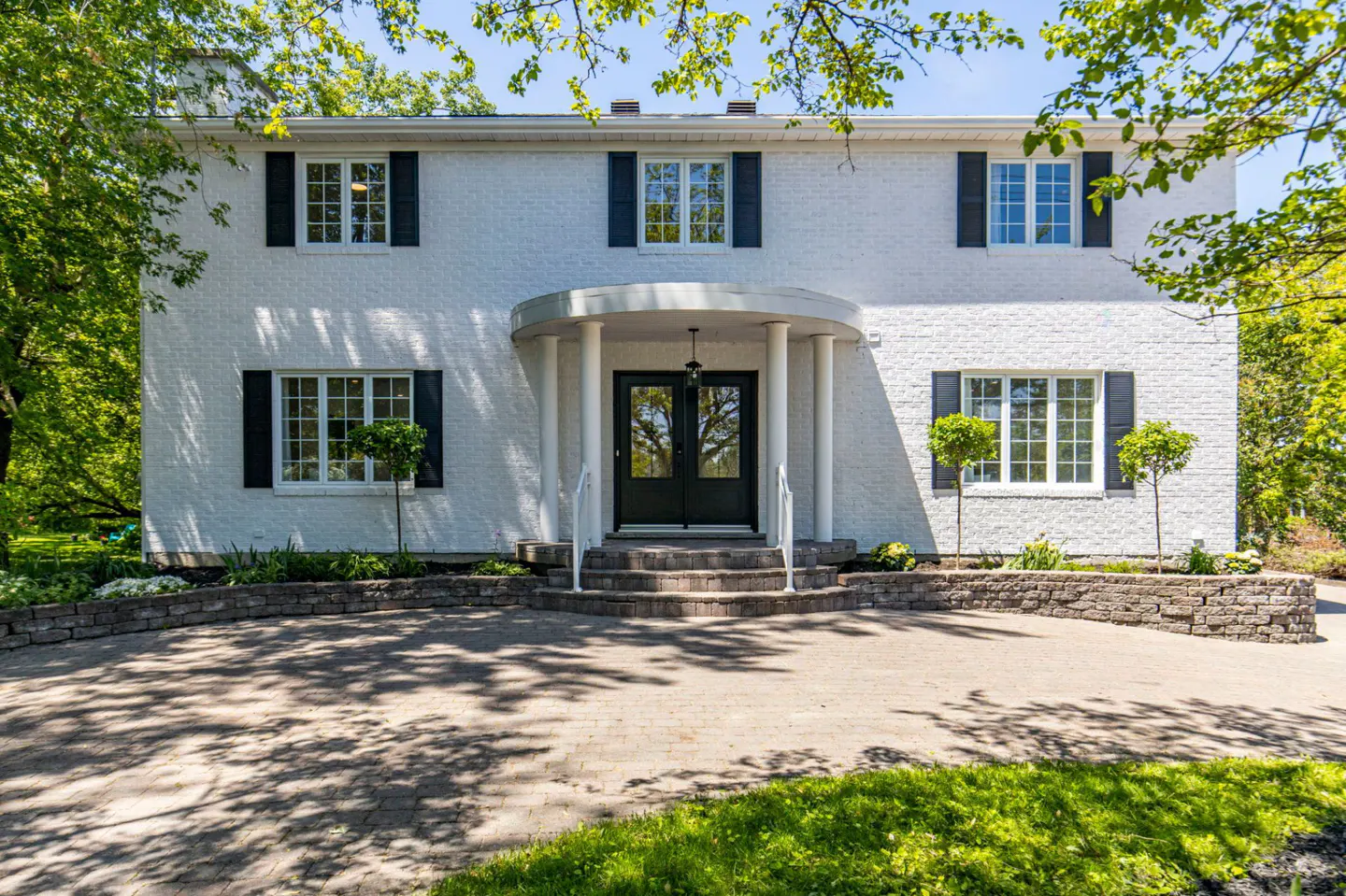 Two-story white brick colonial home with black shutters, a curved front porch, and a paved driveway surrounded by trees.