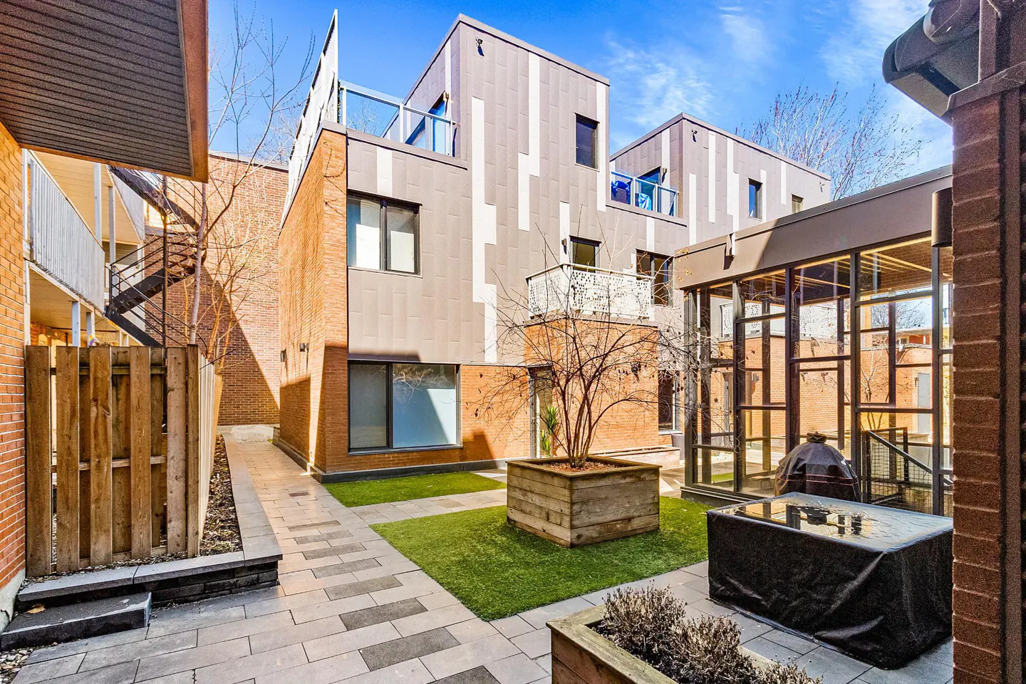 Courtyard view of modern townhouses with brick and gray siding, stone pavers, and small patches of green grass. A bare tree sits in a wooden planter.