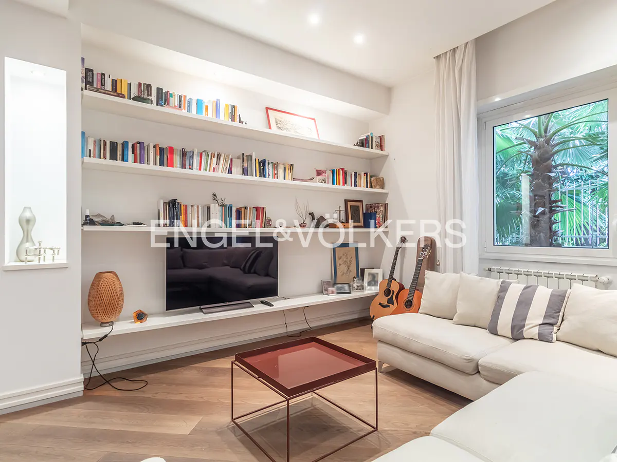 Living room with white shelves filled with books, a TV, guitars, and a white sectional sofa. A red coffee table sits on a wood floor.