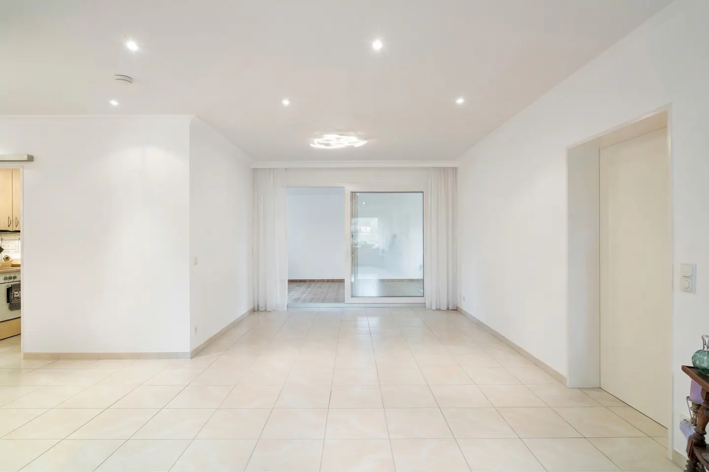 Bright, empty room with white walls, tile floor, and sliding glass doors with sheer white curtains. A glimpse of the kitchen is visible.