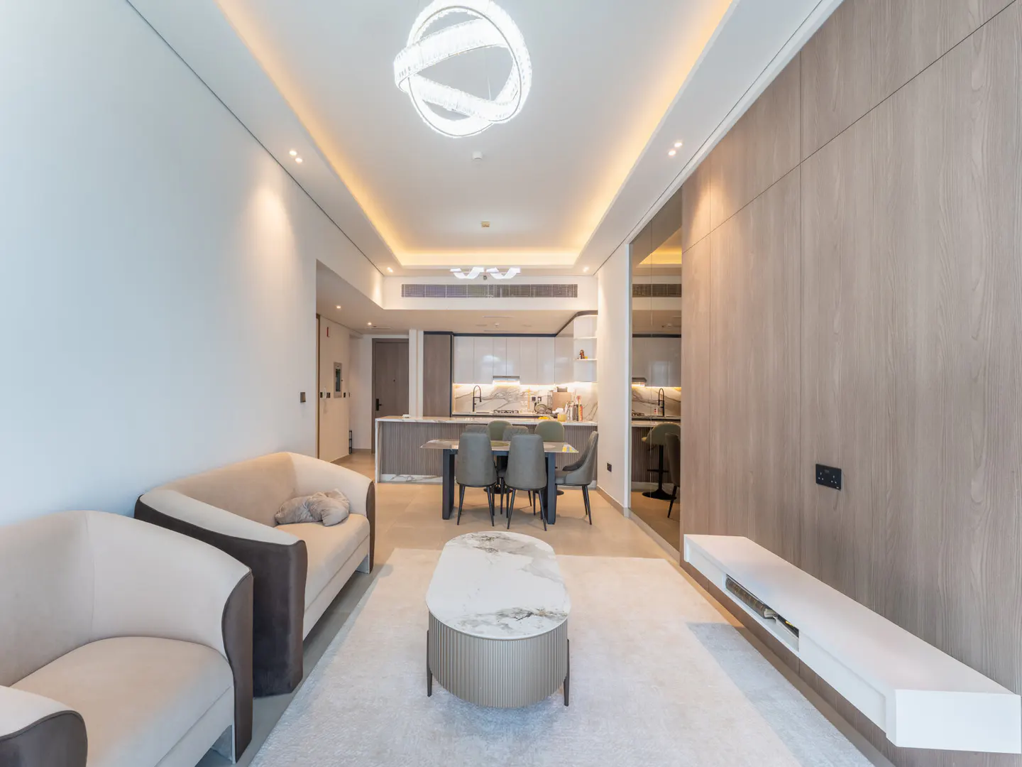 A modern apartment interior with a beige sofa, dining table, and open kitchen. A white rug and a marble-topped coffee table add elegance.