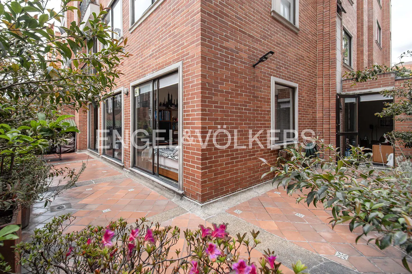 Red brick building exterior with a tiled patio, sliding glass doors, and lush green plants with pink flowers in the front.