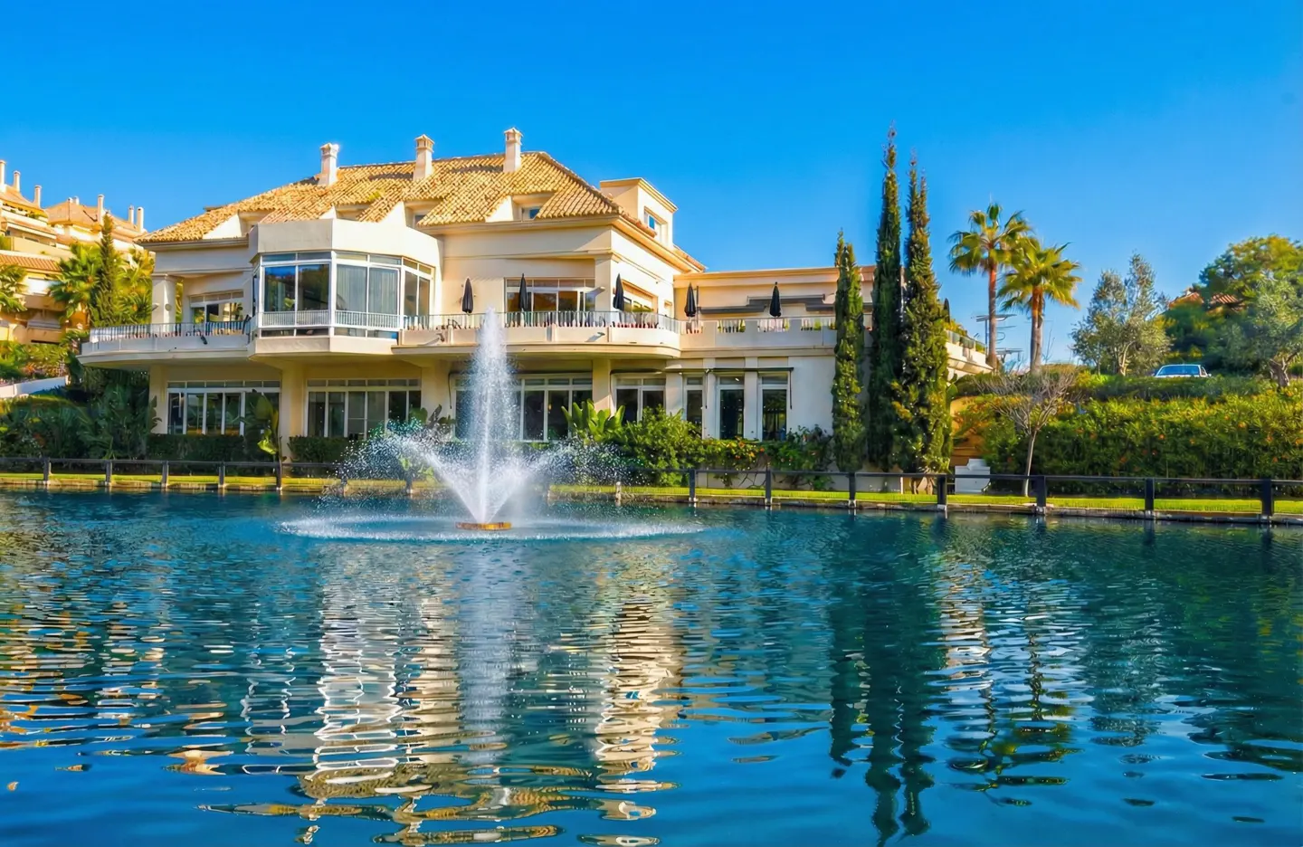 Luxury home with a fountain in a pond. The house is beige with a brown tile roof. The sky is blue.