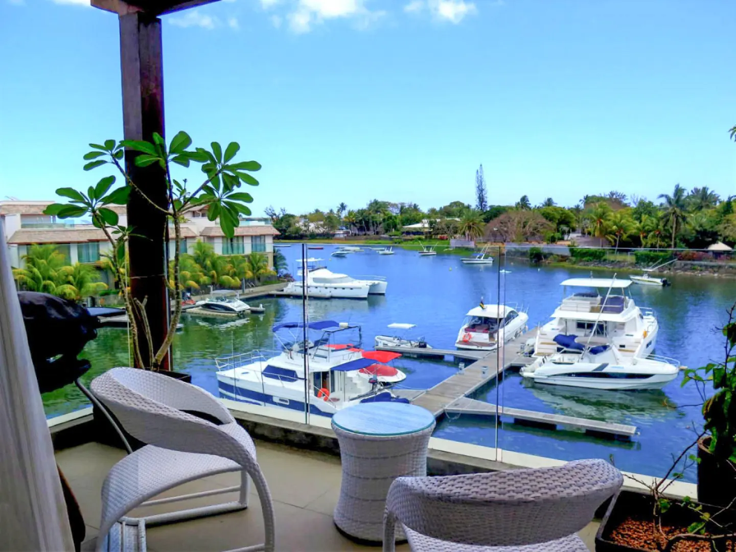 Balcony view of a marina with boats docked in blue water under a sunny sky. White wicker chairs and a table are on the balcony.