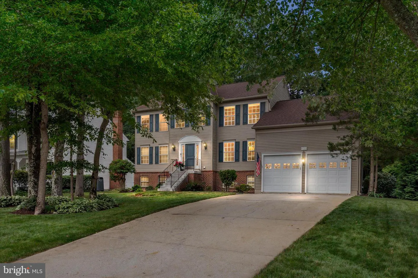 Two-story beige suburban home with a two-car garage, concrete driveway, and lush green trees in a landscaped front yard.