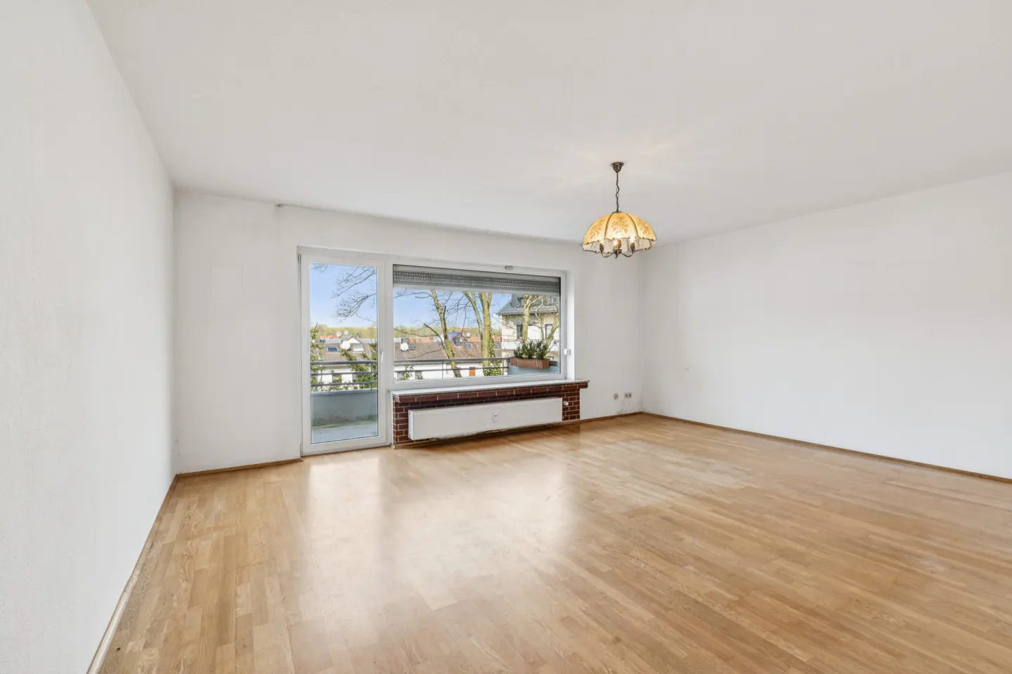 Bright, empty room with hardwood floors, white walls, and a large window overlooking a balcony and neighborhood. A vintage chandelier hangs from the ceiling.