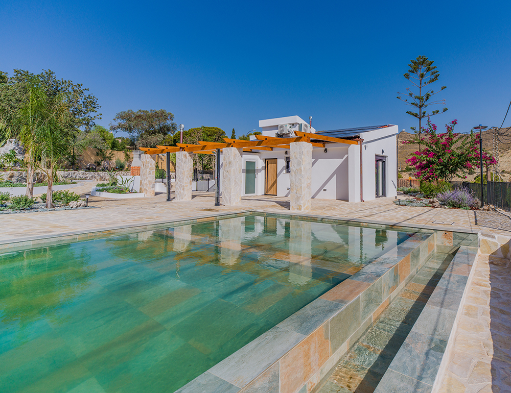 Modern white villa with a wooden pergola and swimming pool in a sunny, landscaped garden under a clear blue sky.