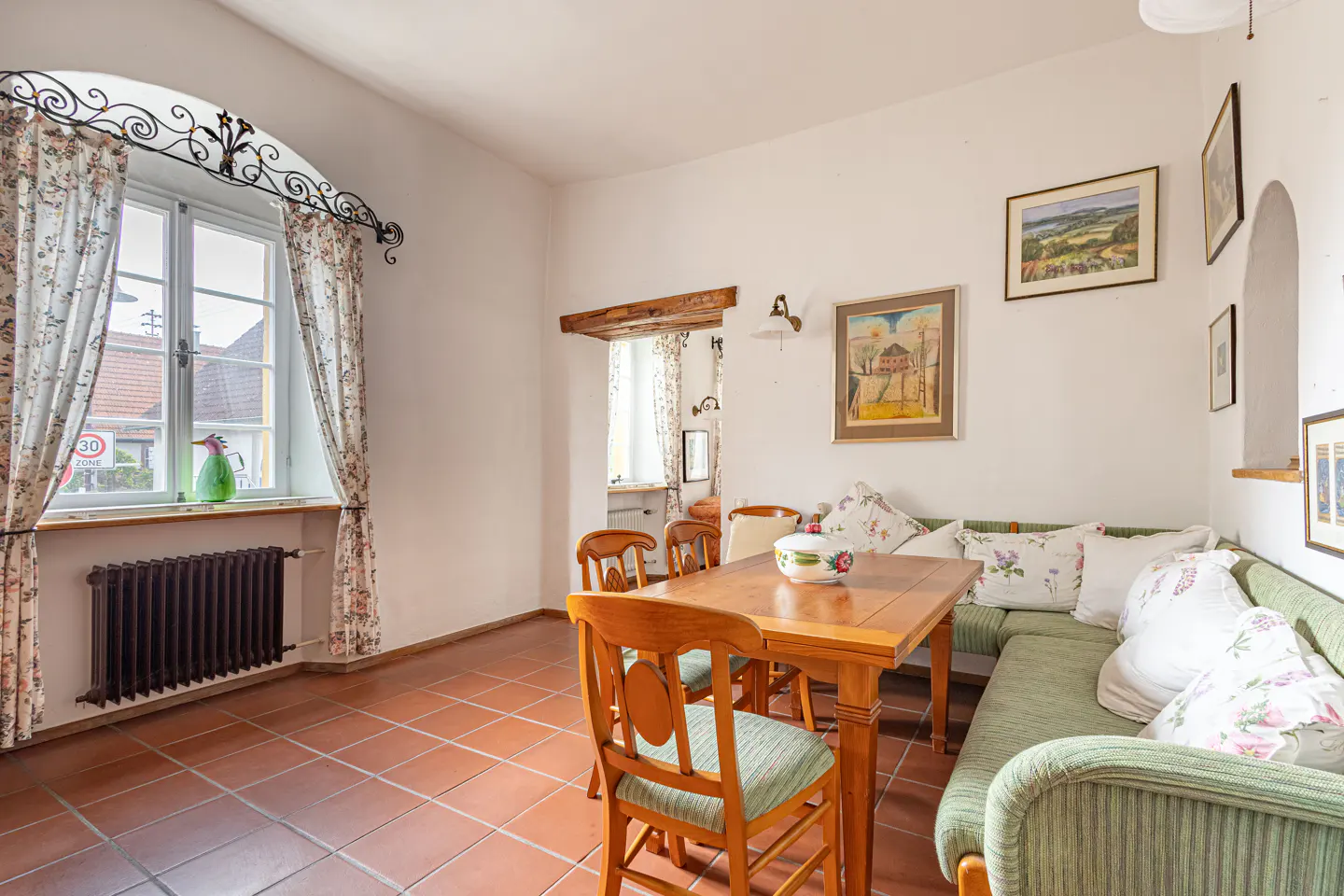 Bright dining room with terracotta tile floor, wooden table and chairs, and a green cushioned corner bench. Floral curtains frame a window with a black iron decorative piece.