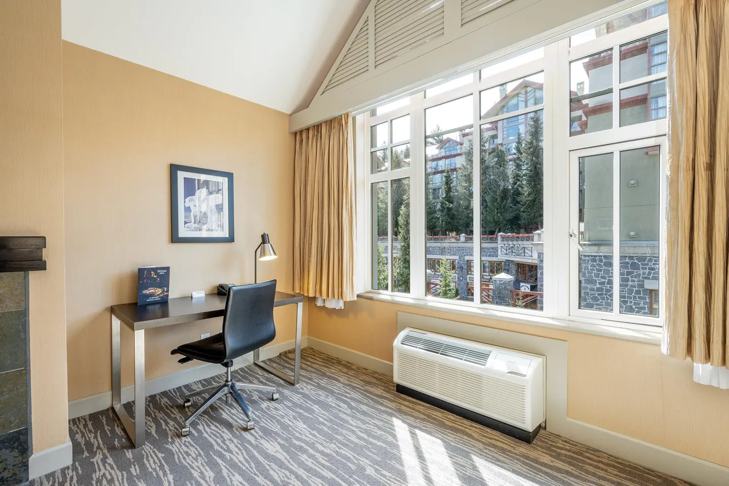 Hotel room interior with a desk, chair, and large window overlooking trees and buildings. Beige walls and patterned carpet.
