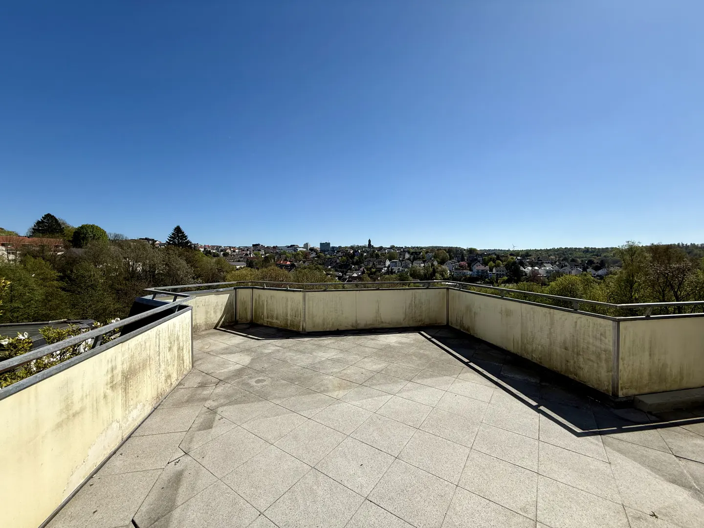 Wide shot of a large, empty, tiled balcony with a city view under a clear blue sky.