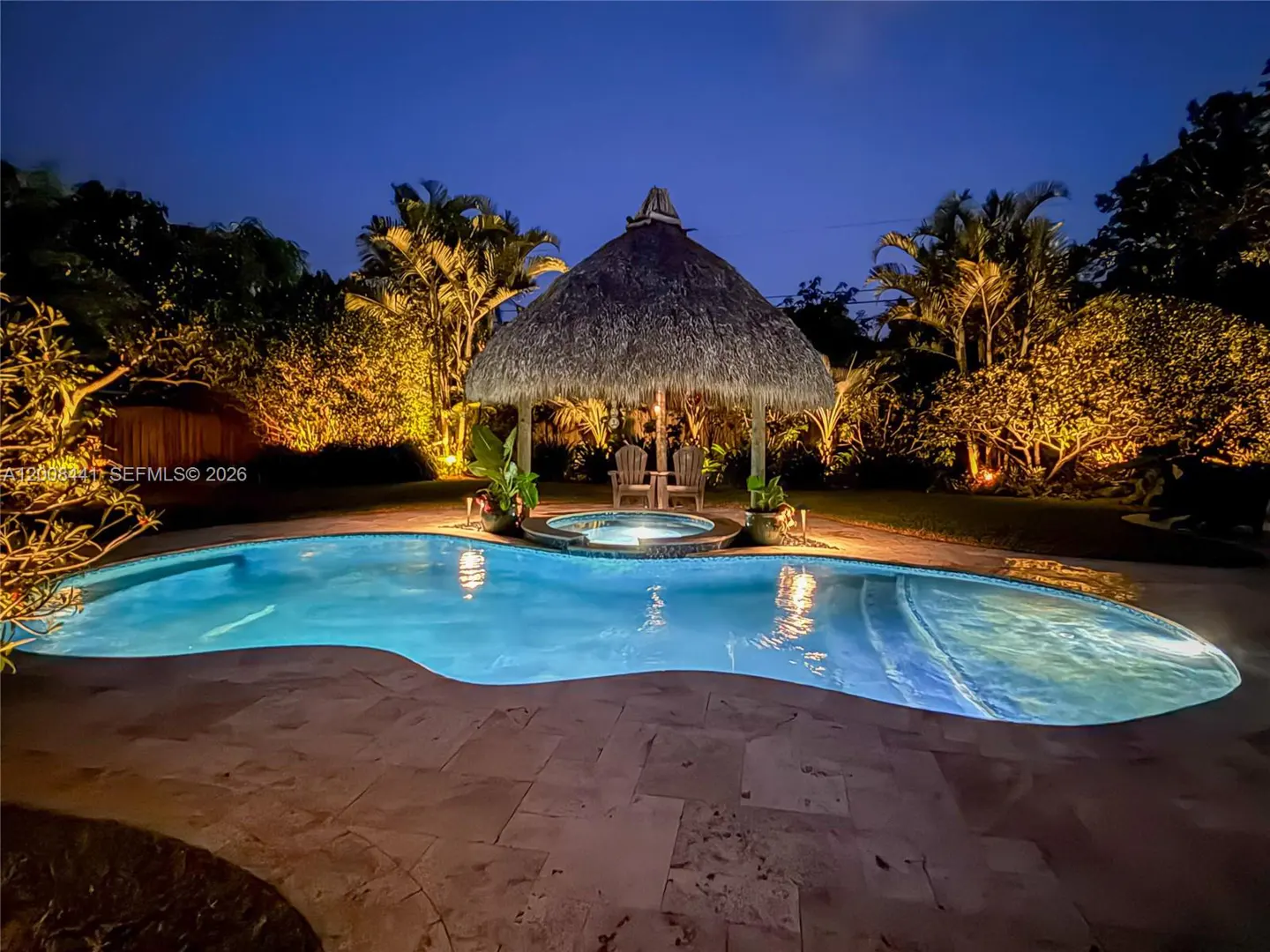 Night view of a backyard pool with a thatched-roof gazebo, two chairs, and lush landscaping illuminated by warm lighting.